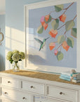 Colorful painting of a hummingbird hovering near pink and coral blossoms against a soft blue background.